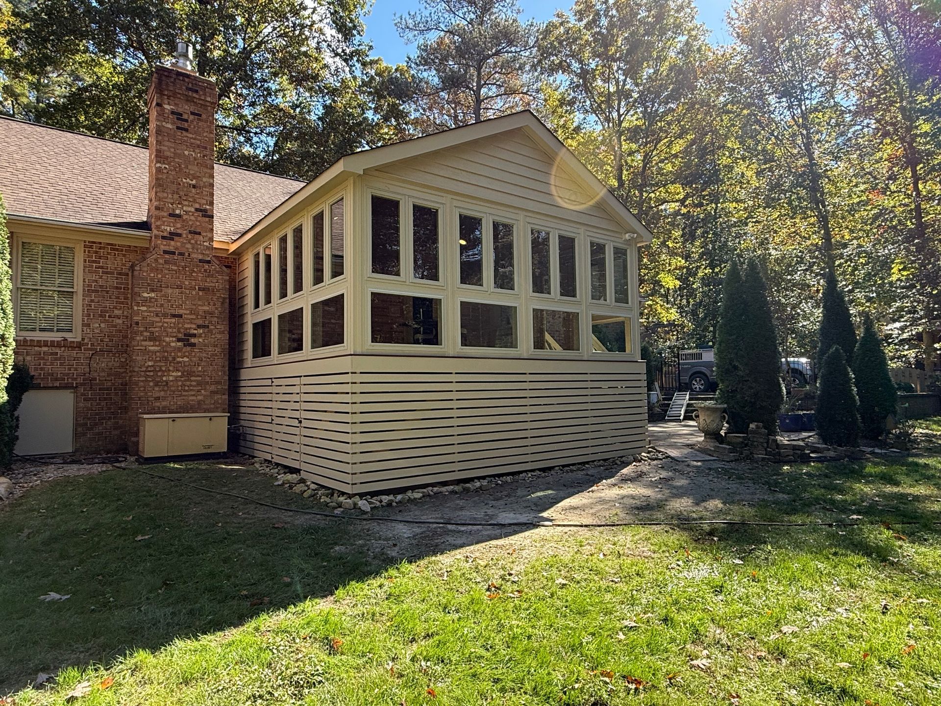 Cream sunroom addition on a brick house, with many windows, surrounded by trees and a grassy yard.