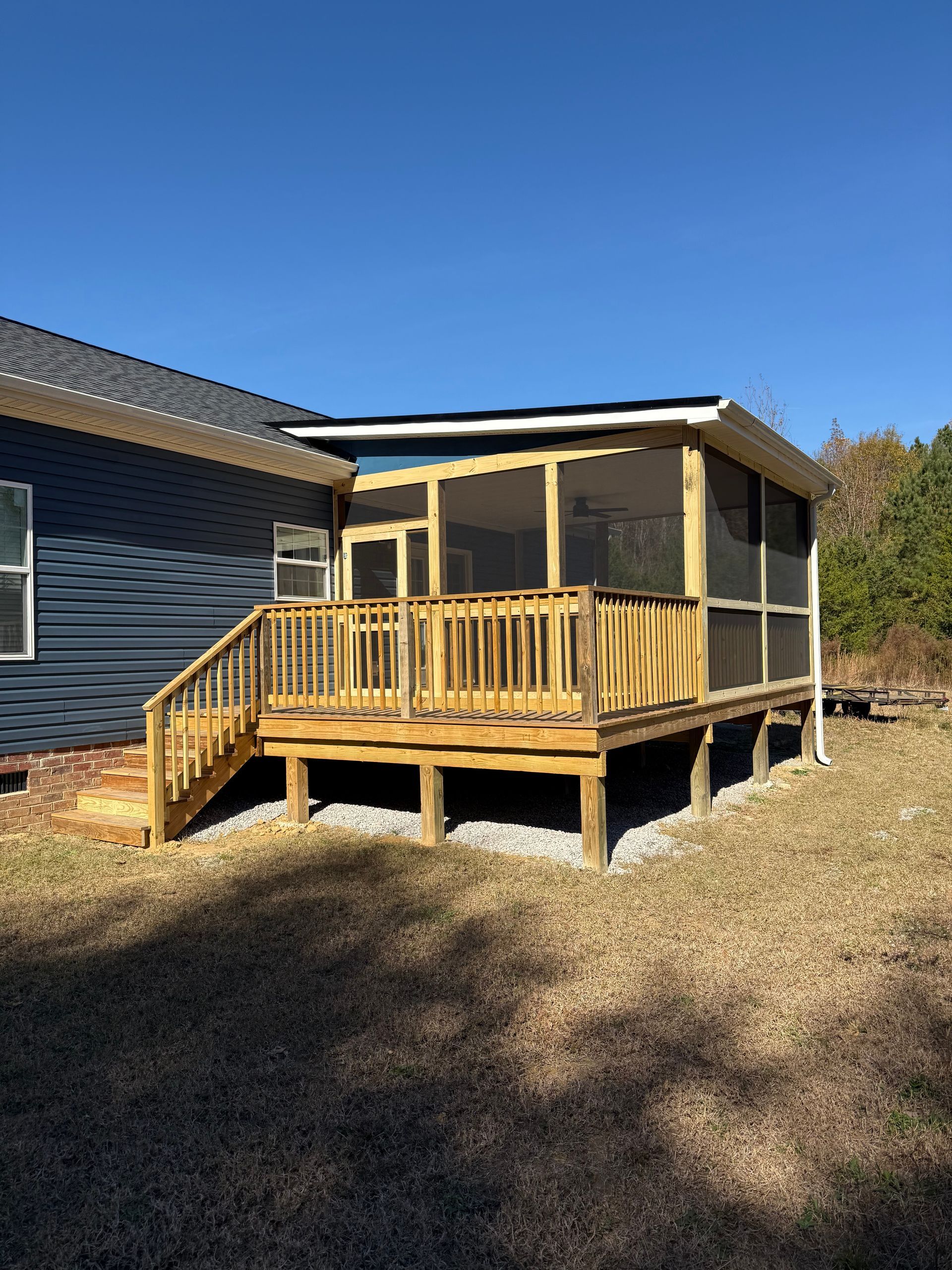 Wooden screened porch deck attached to a house in a sunny yard with bare ground and blue sky
