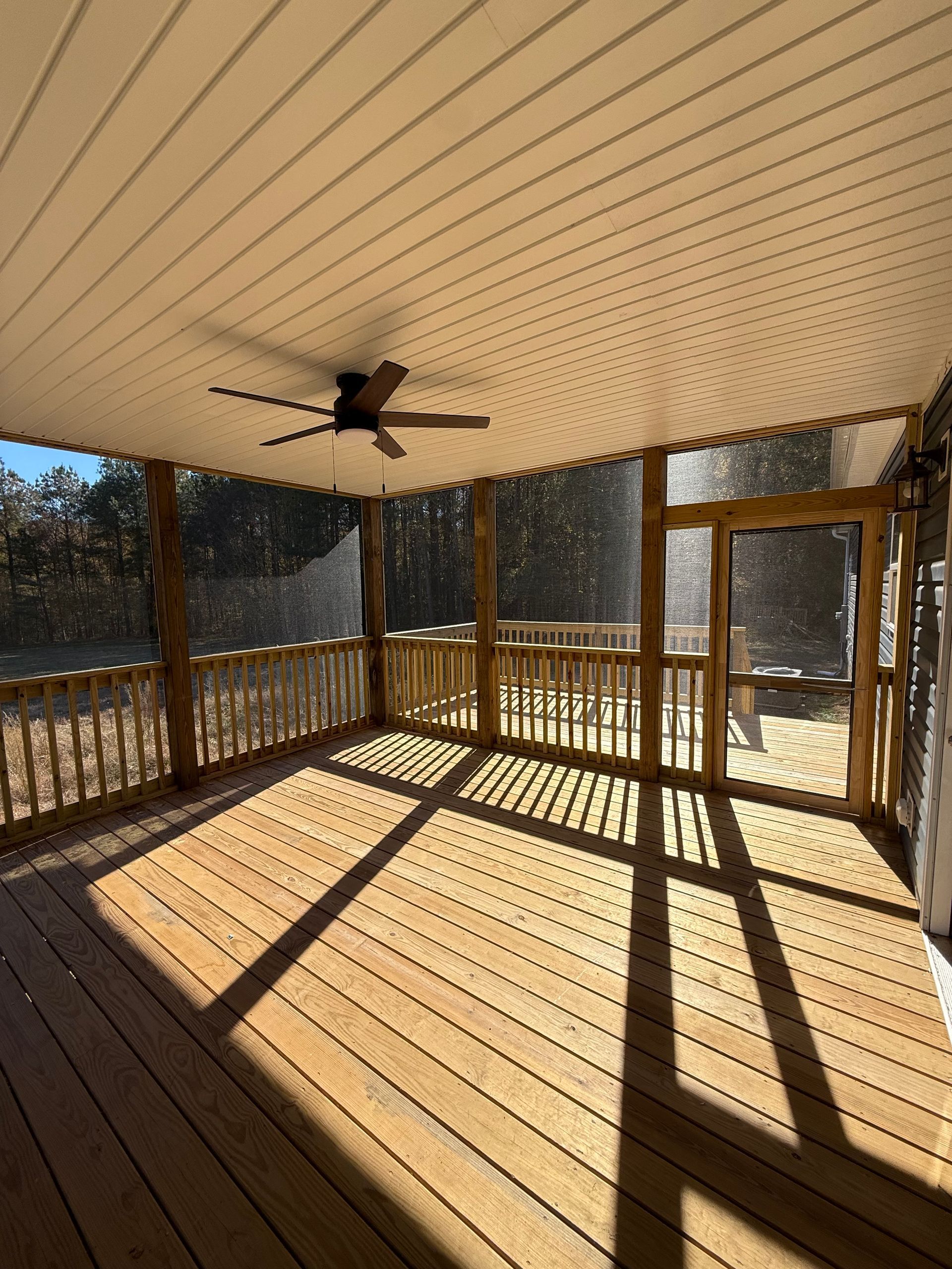 Covered wooden deck with railing, screened walls, and a ceiling fan in bright sunlight