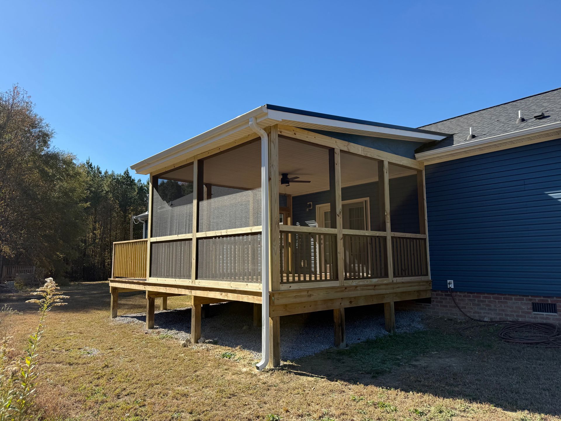 Covered wooden porch with railings attached to a blue house in a sunny yard