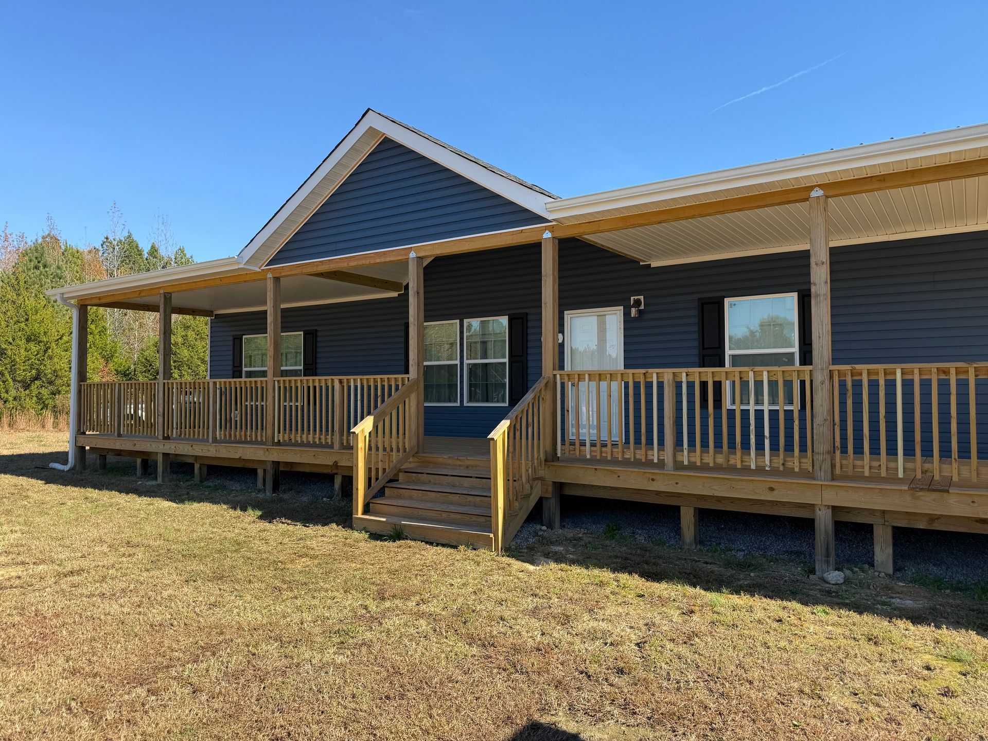 Single-story house with a front porch and steps, dark siding, and a grassy yard under a clear blue sky