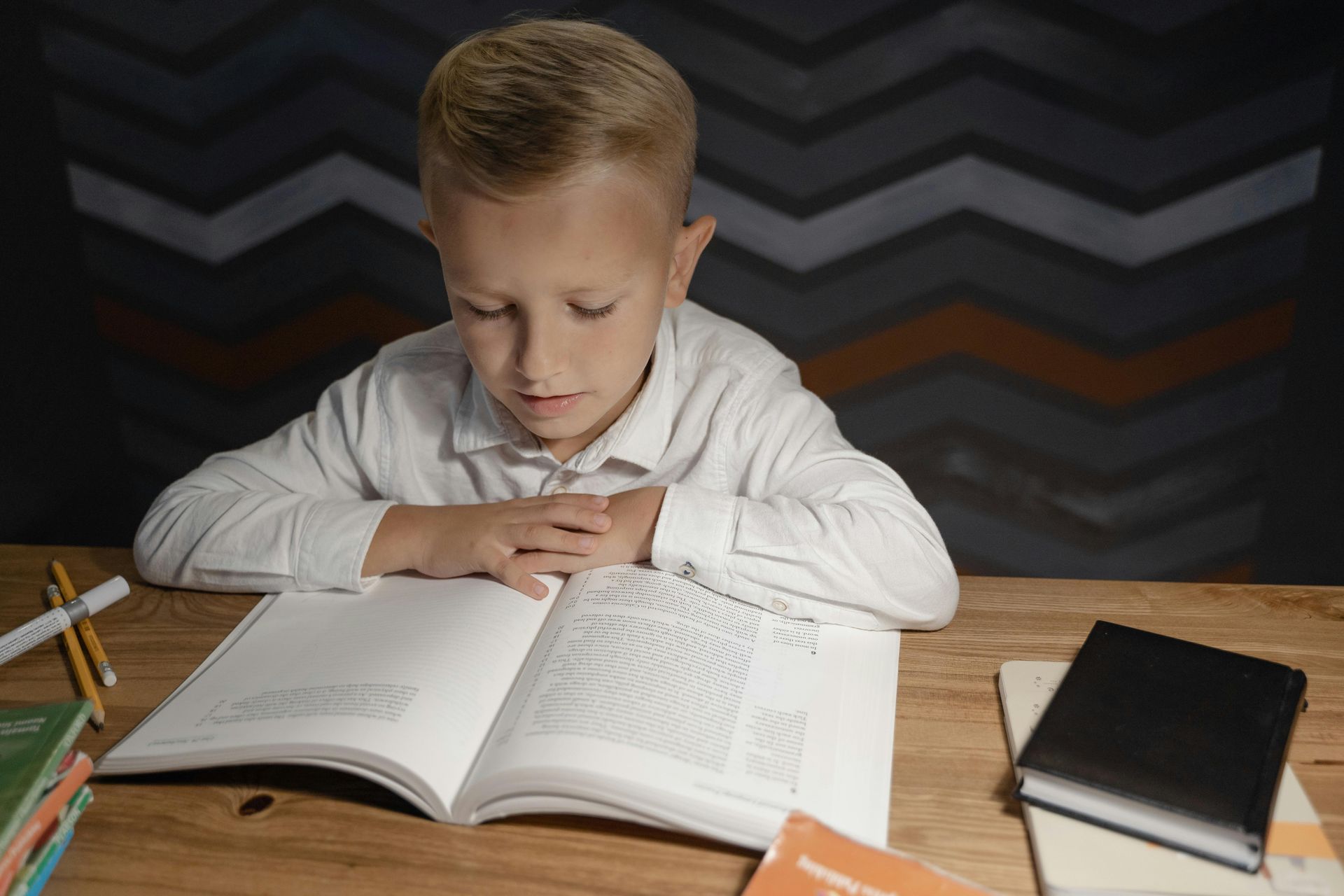 Child reading an open book at a wooden desk, with notebooks and a black book nearby.