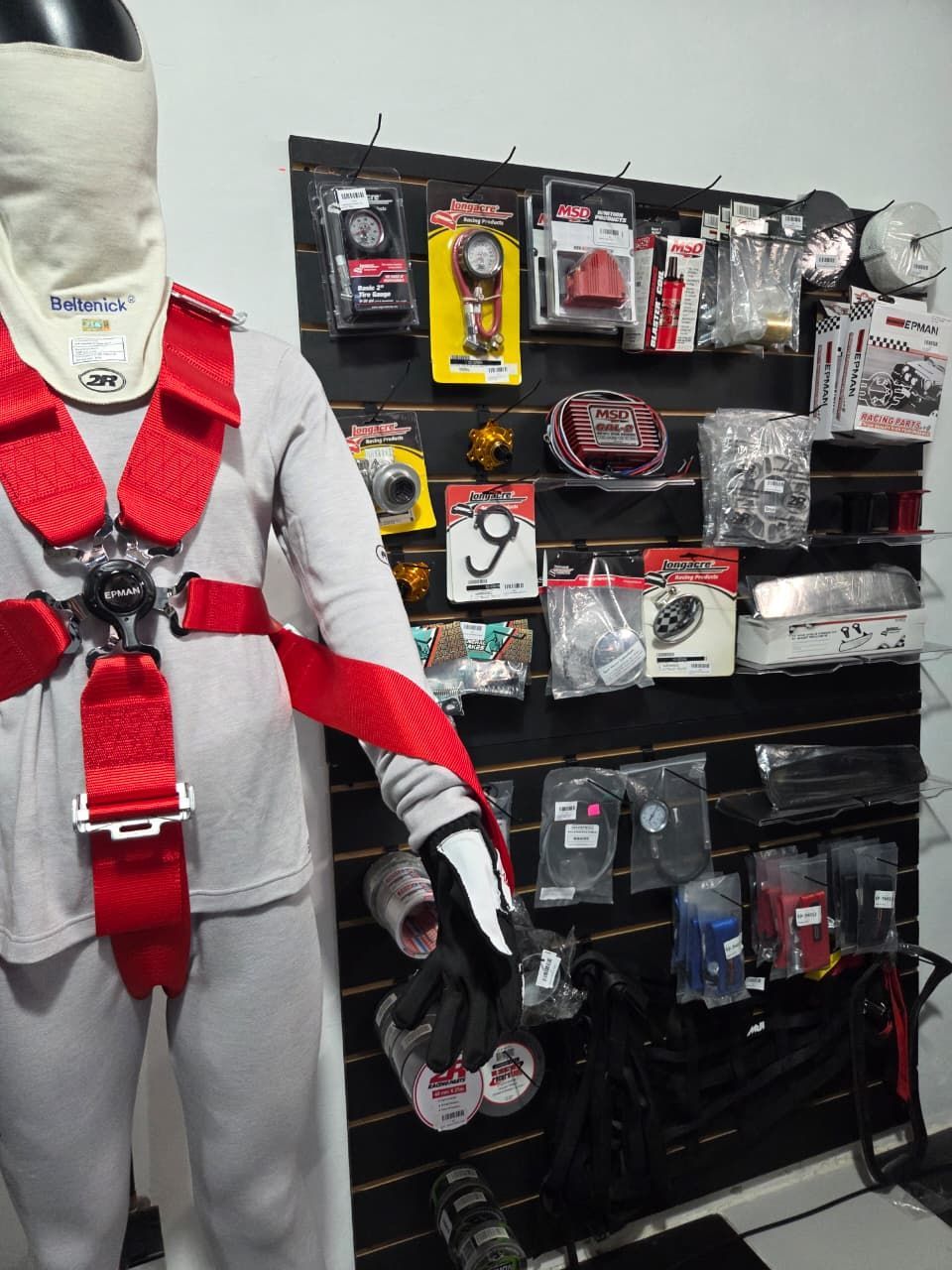 Maniquí blanco con arnés de seguridad rojo junto a una pared llena de equipos y accesorios de seguridad empaquetados.