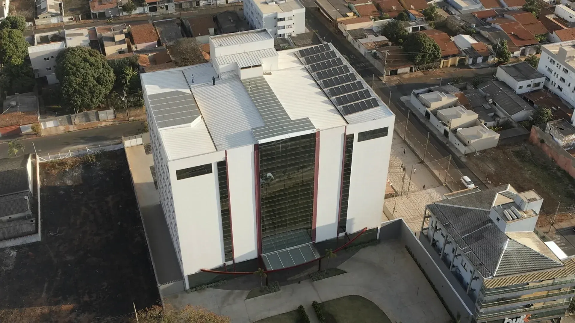 Vista aérea de um edifício moderno branco com painéis solares no telhado; entrada com vidro vermelho e transparente.