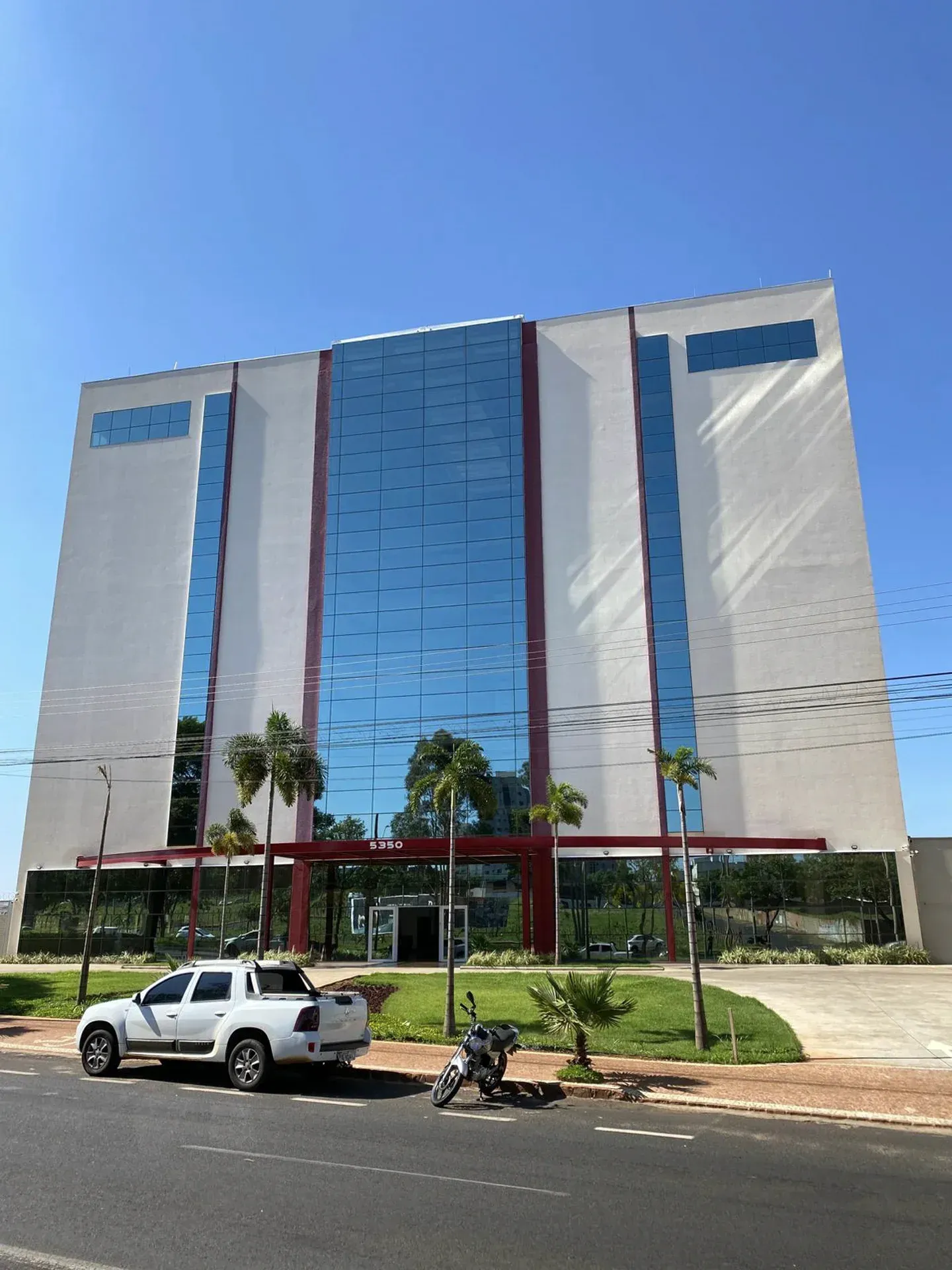 Prédio moderno de vários andares com fachada de vidro e exterior branco, céu azul, carro branco estacionado em frente.