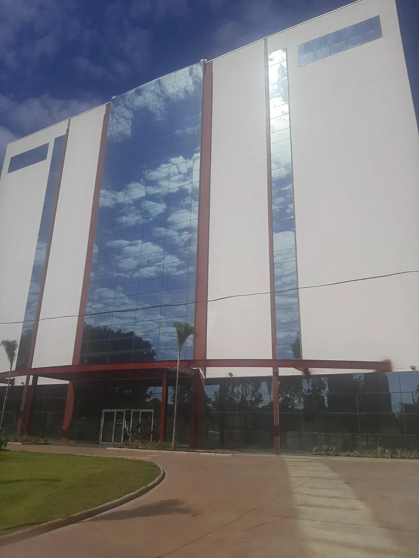 Prédio branco e de vidro que reflete o céu azul, com detalhes em vermelho na entrada.