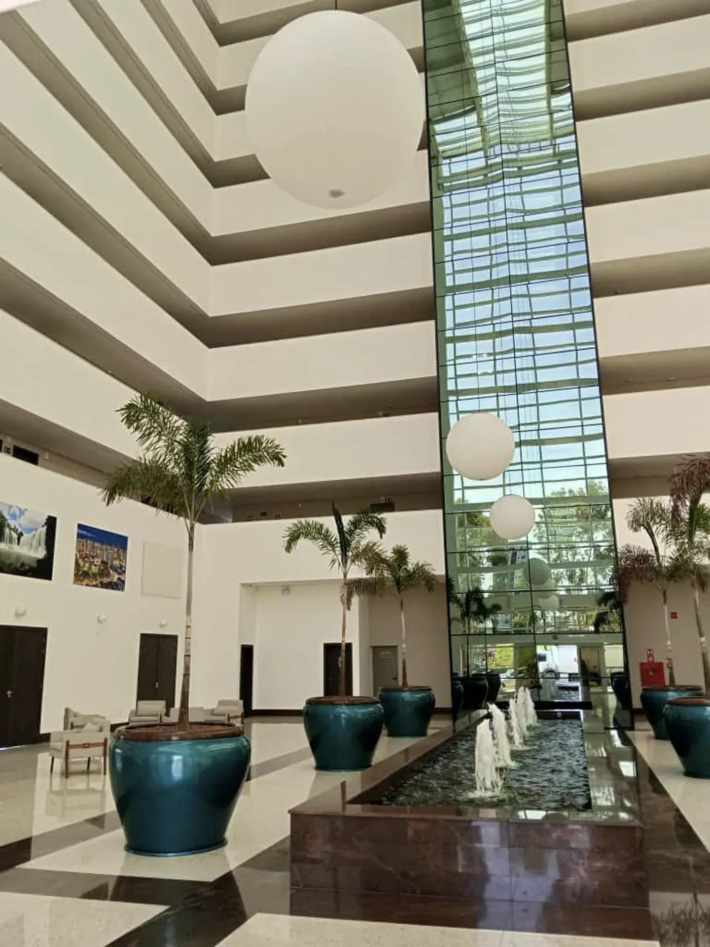 Hall de entrada com arquitetura em vários níveis, grandes globos de luz, plantas de interior, fonte de água e elevador.