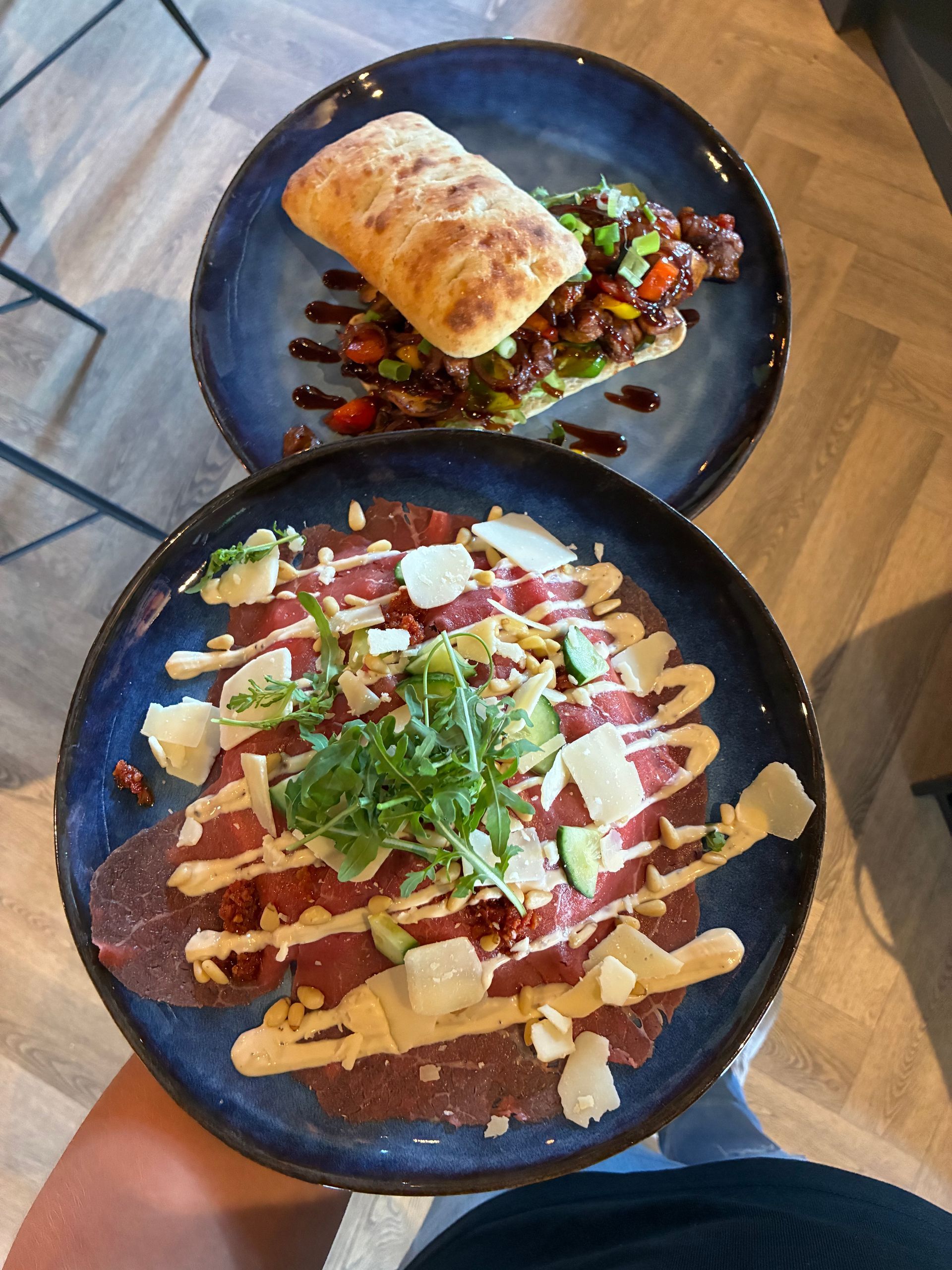 Twee borden met eten op blauwe borden. De ene met een sandwich, de andere met rundercarpaccio in de hand.
