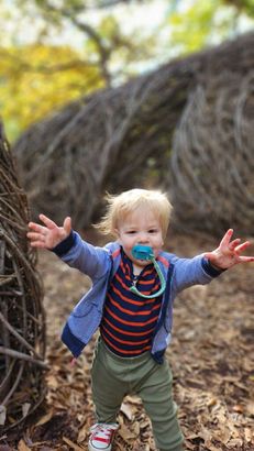 A little boy with a pacifier in his mouth is standing in the woods with his arms outstretched.