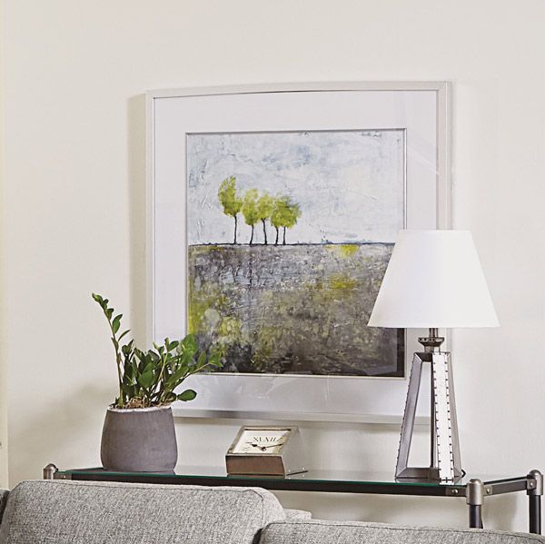 Framed abstract landscape above a console table with a plant, lamp, books, and a gray sofa arm