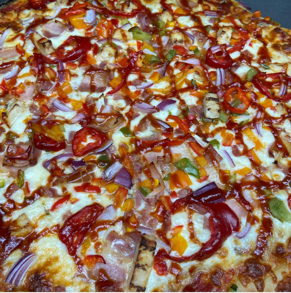 A pizza topped with chicken, red onions, mushrooms, green peppers, and BBQ sauce — all served on a wooden cutting board. Pineapple and hot peppers available upon request.