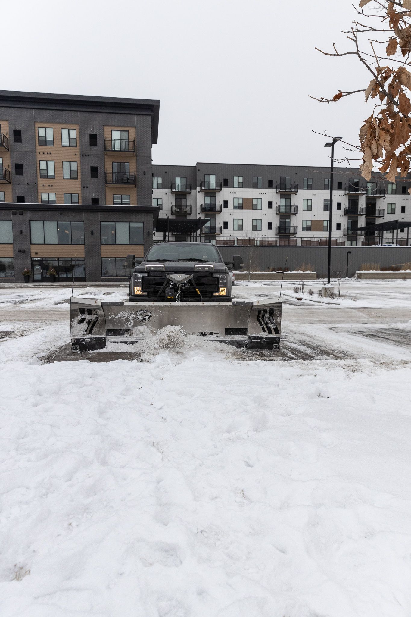 A snow plow is clearing snow from a parking lot in front of a building.