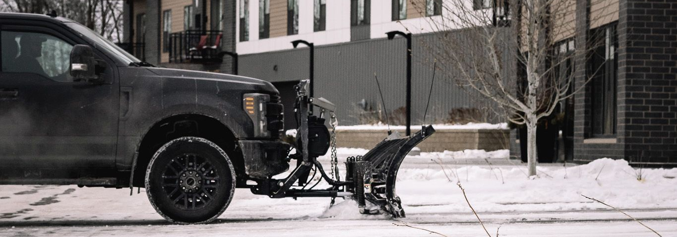 A dark truck with a snowplow on a snowy street in front of a building.