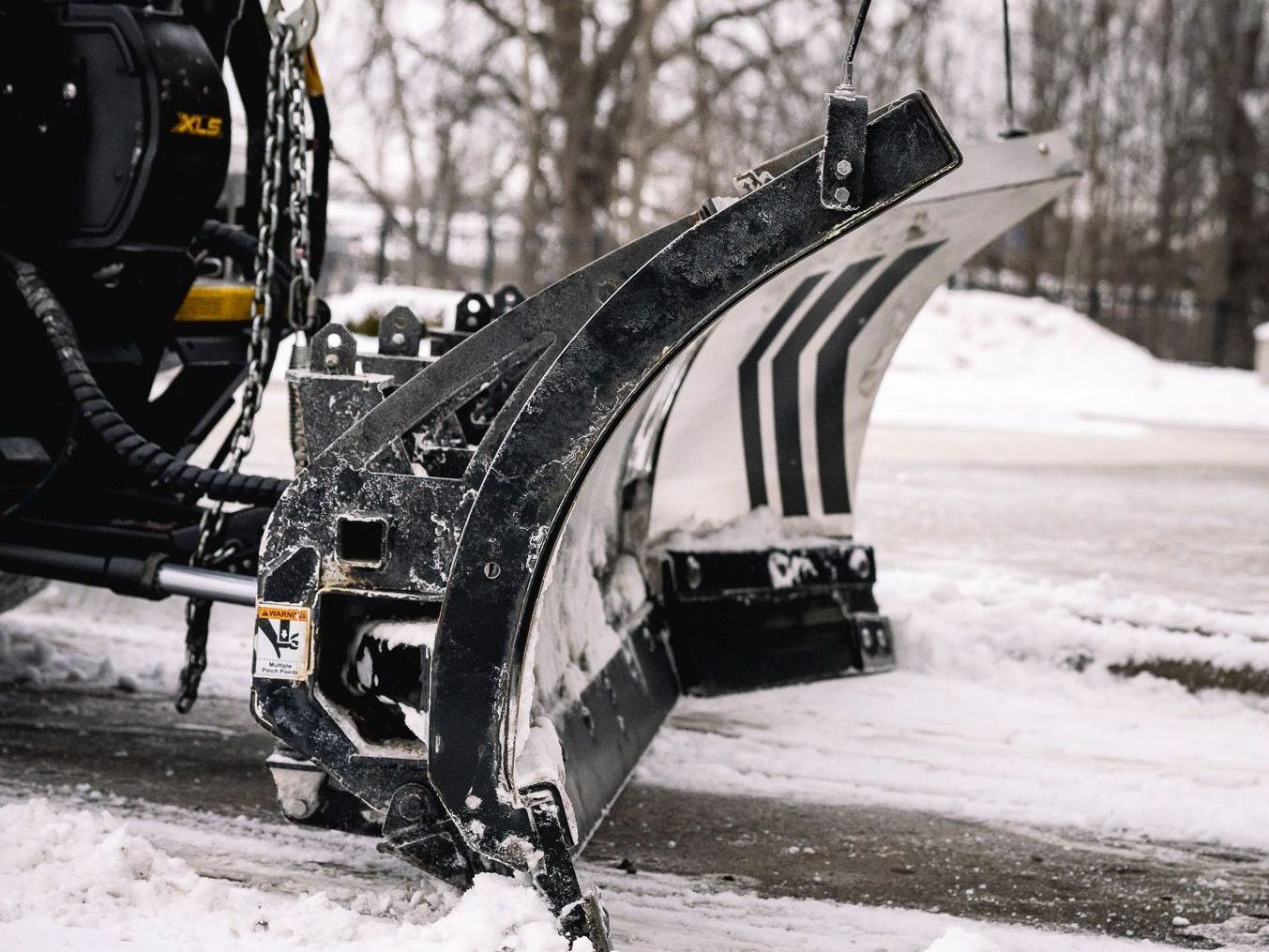 Snowplow blade angled down, clearing snow from road. Gray blade with black frame, chains, and snow.