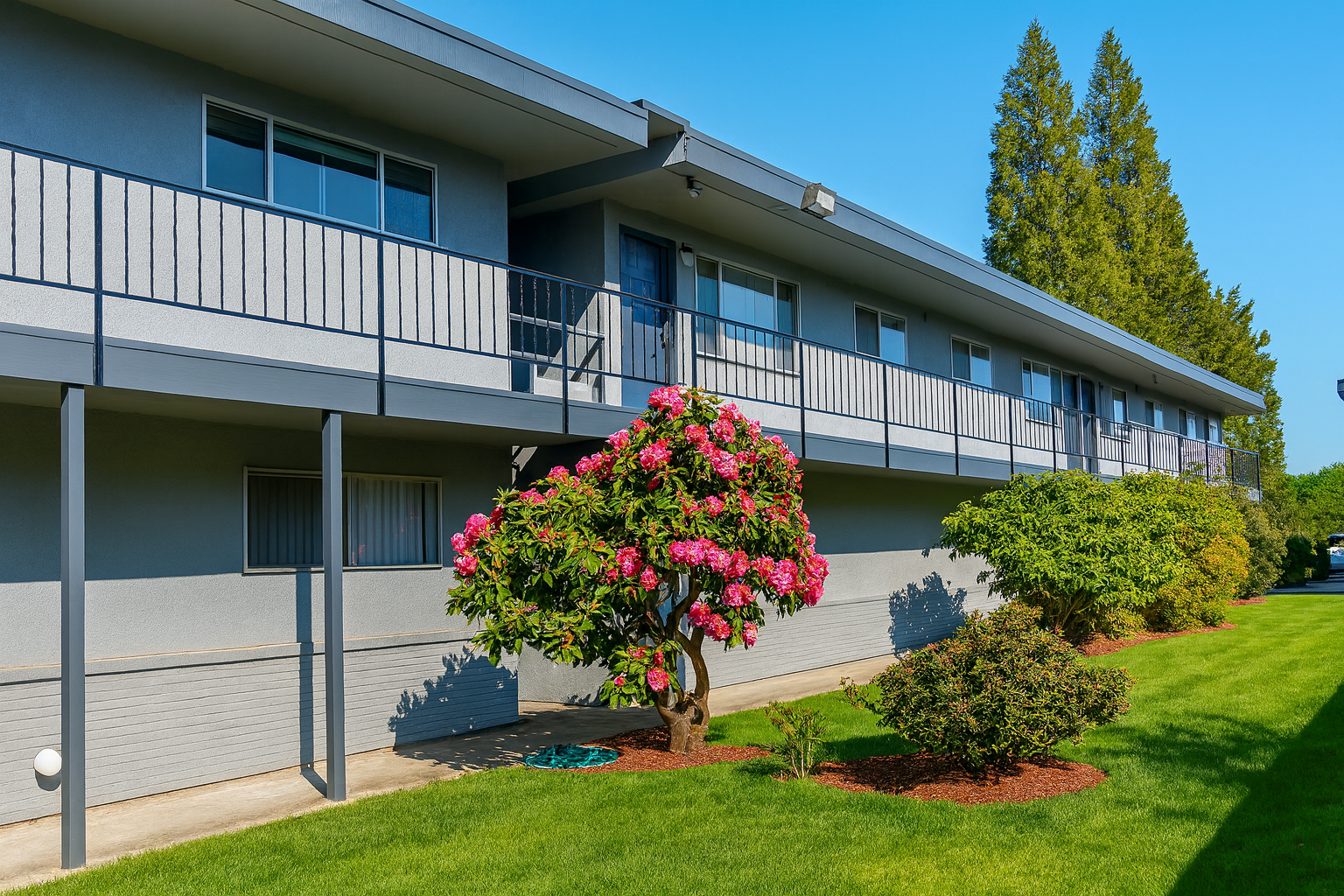 Two-story apartment building with balconies, gray exterior, green lawn, and flowering bush.