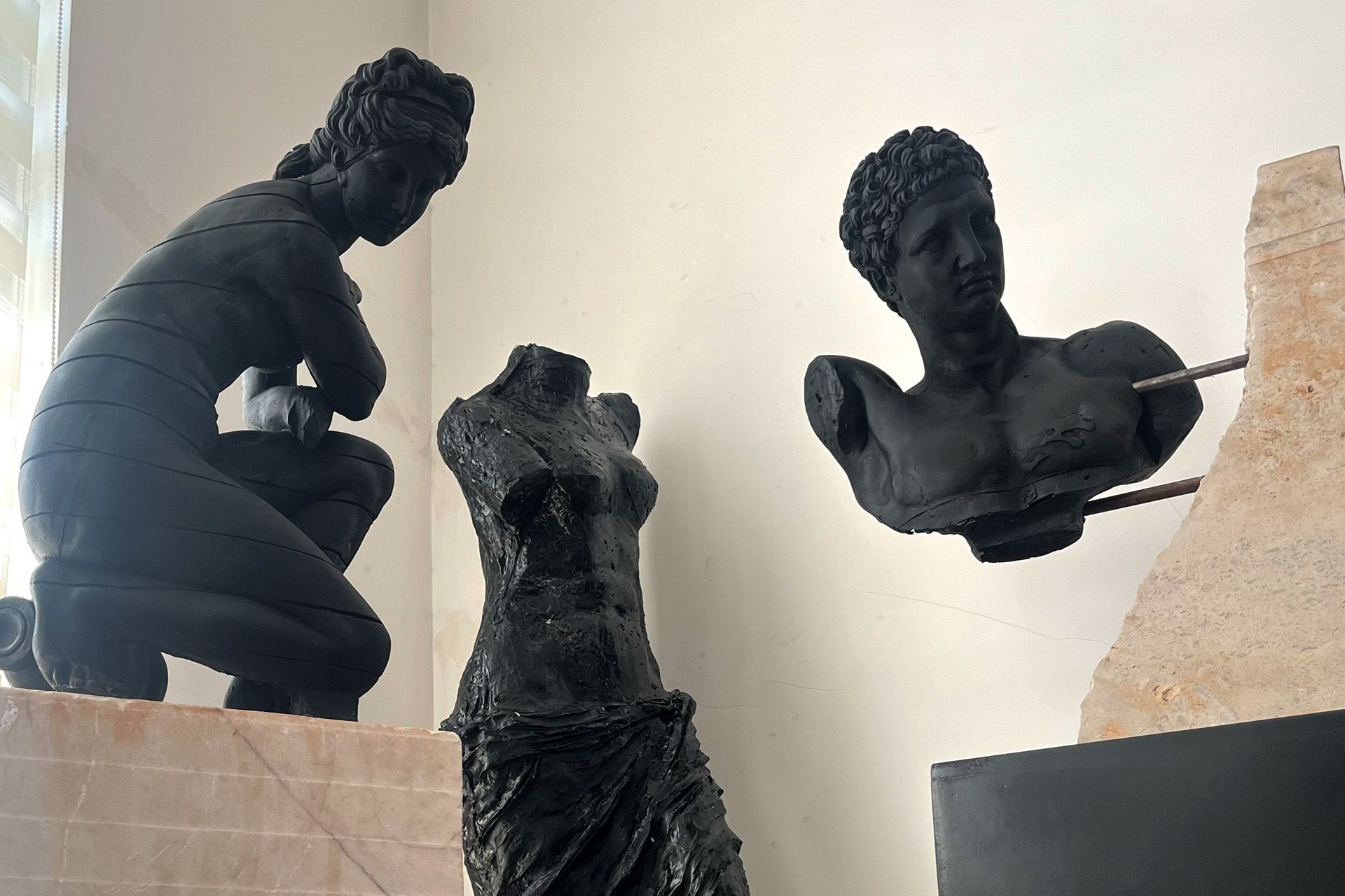 Esculturas clásicas negras: una figura arrodillada, un torso y un busto pegados a una pared, todo en una habitación luminosa.