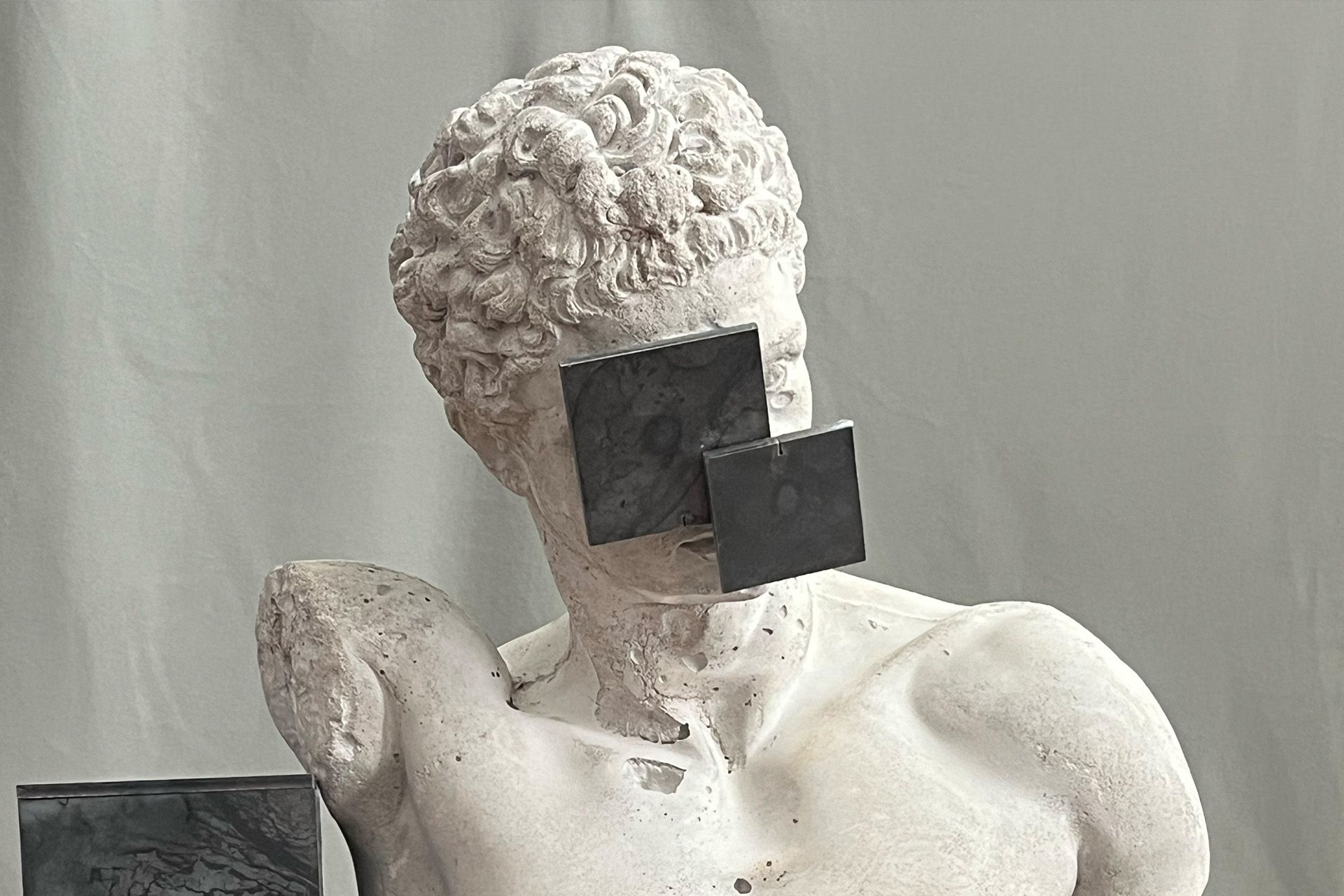 Busto de estatua de mármol con dos cuadrados negros que cubren el rostro, sobre un fondo gris claro.