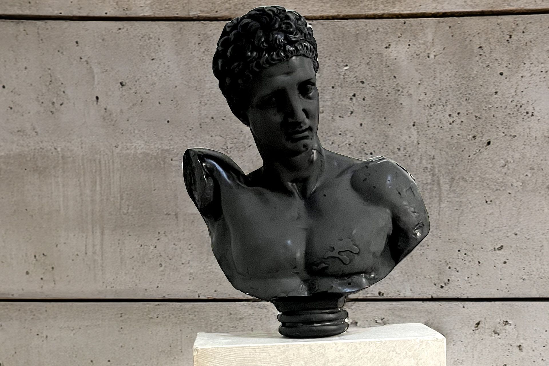 Busto negro de un hombre sobre un pedestal, apoyado contra una pared gris que parece hormigón.