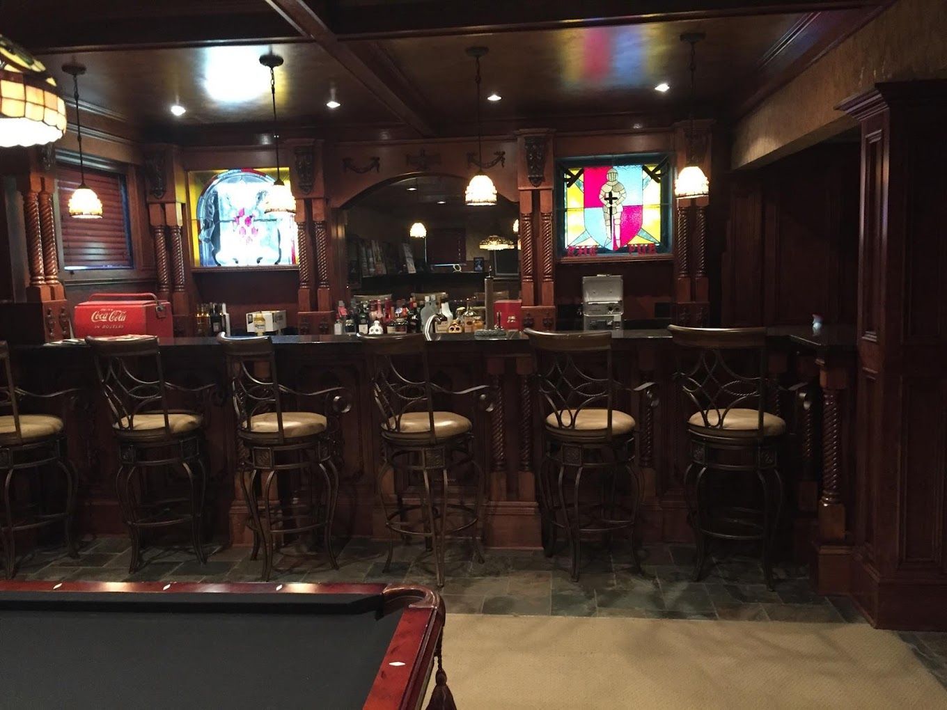 A wood-paneled bar with ornate bar stools, two TVs, and a pool table in a dimly lit basement.