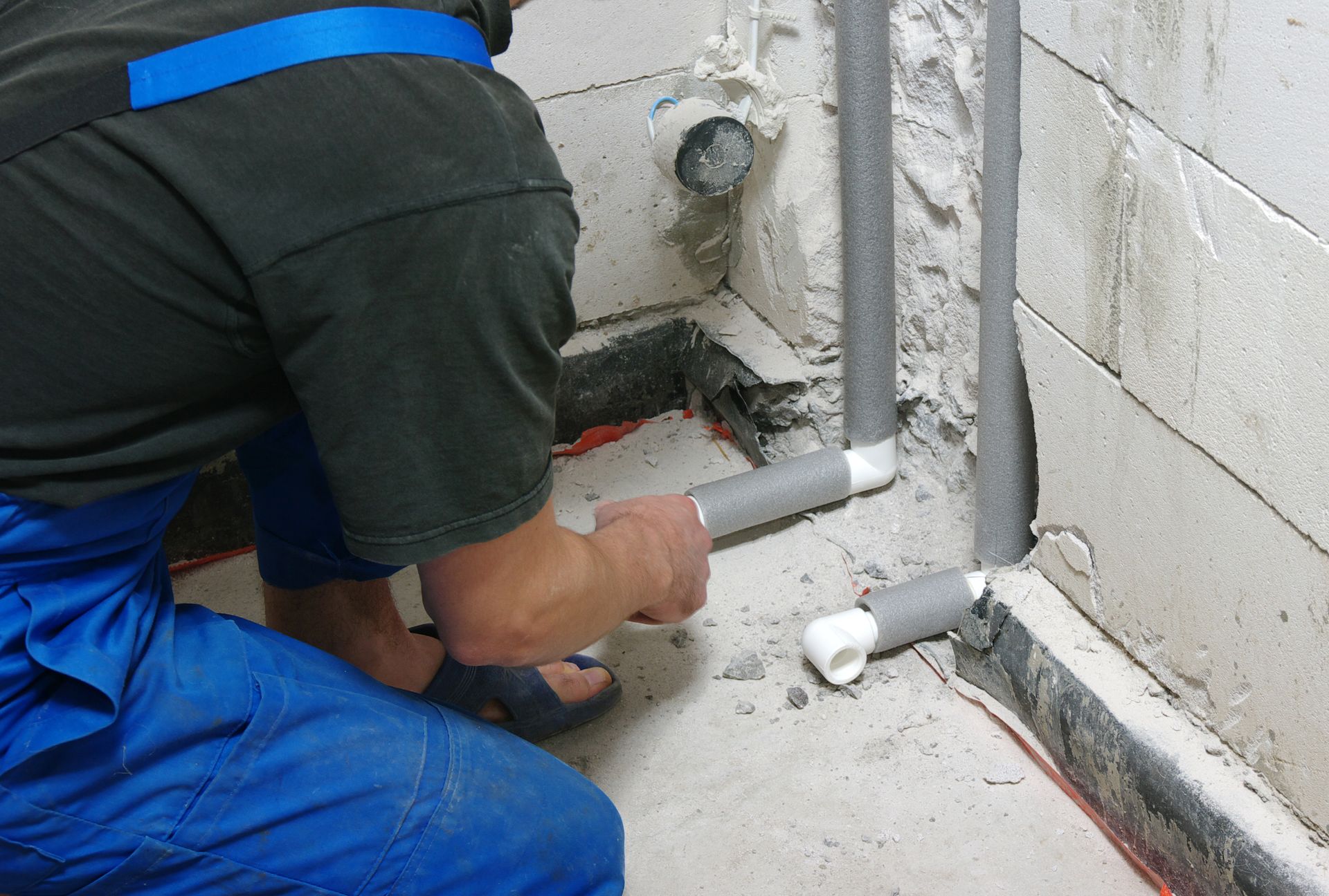 A person installs pipes, wearing blue overalls, in a concrete corner.