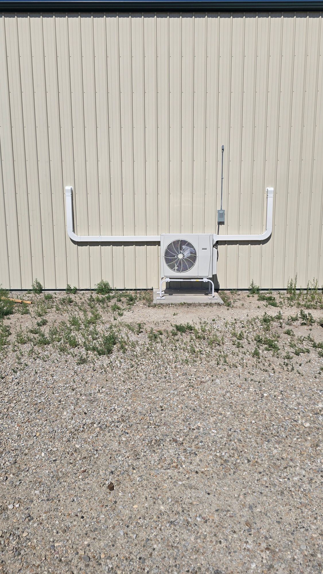 Air conditioning unit on a concrete base, with white pipes extending outwards on a gravel surface.