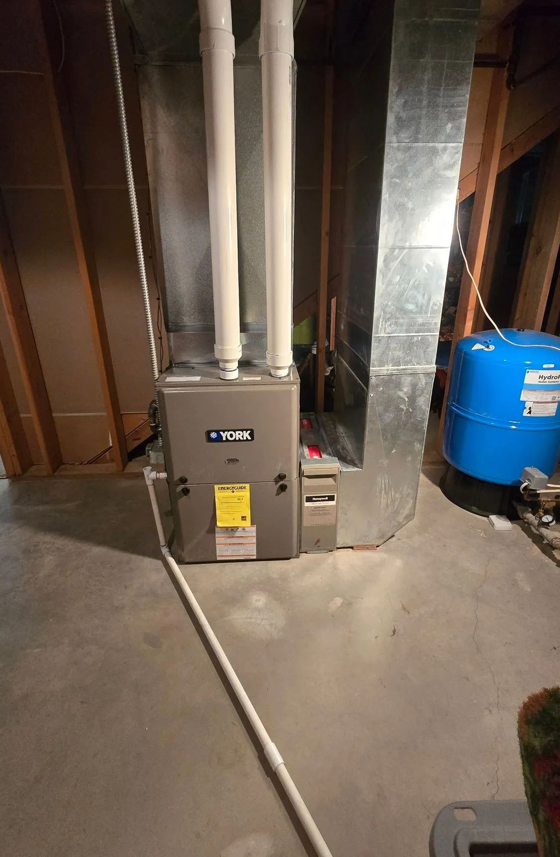 Basement furnace with white ducts beside a blue water heater and exposed framing.