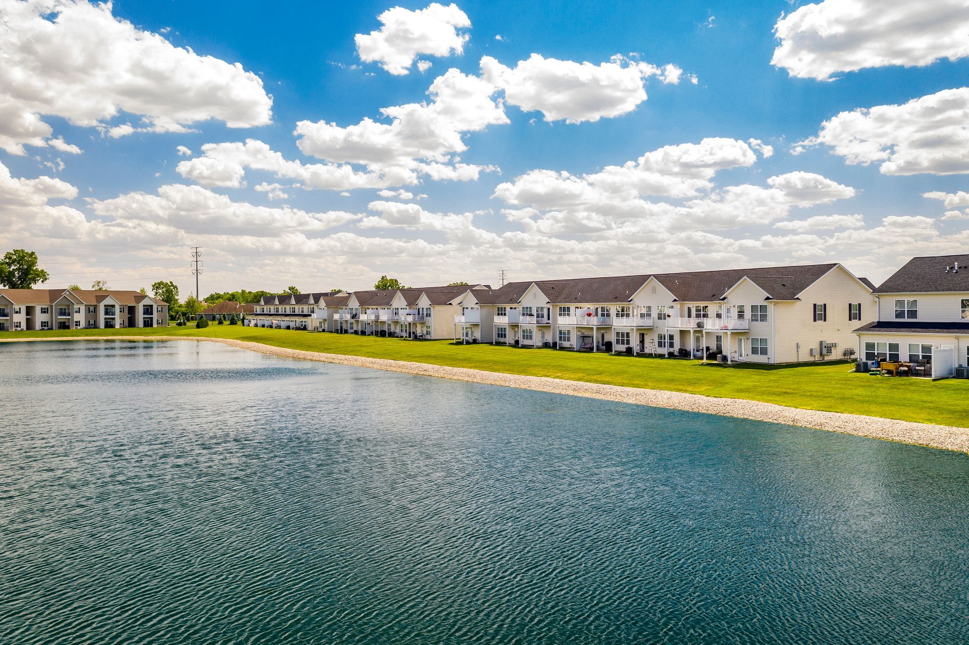 Lakeside row of beige townhomes under a blue sky with fluffy clouds.