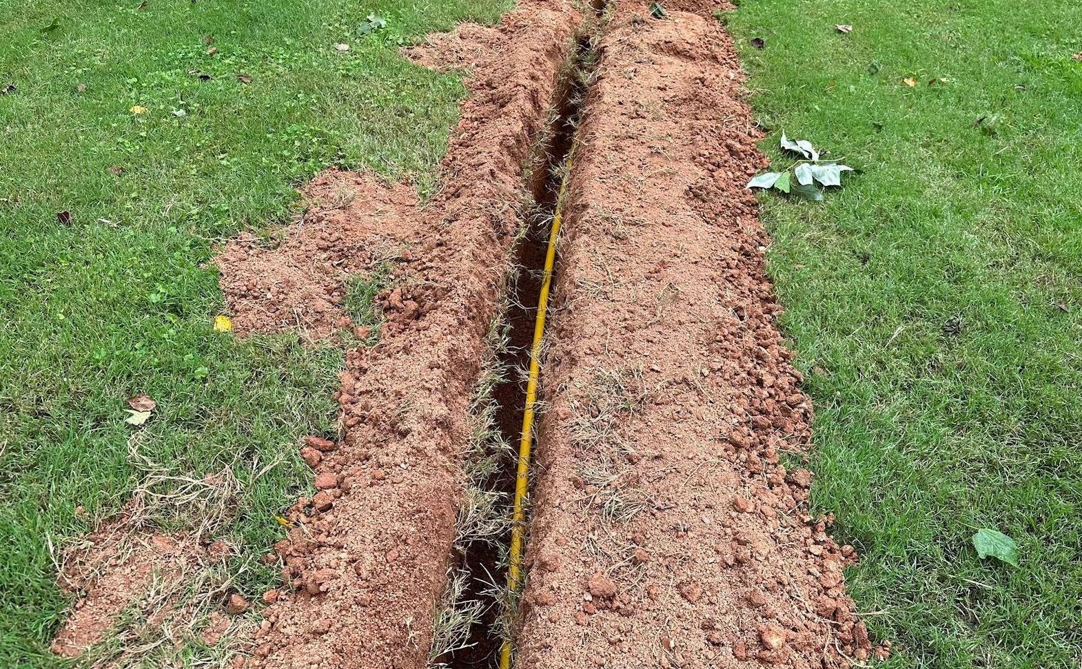 A narrow trench in grass, containing a yellow pipe. Dirt piles on either side.