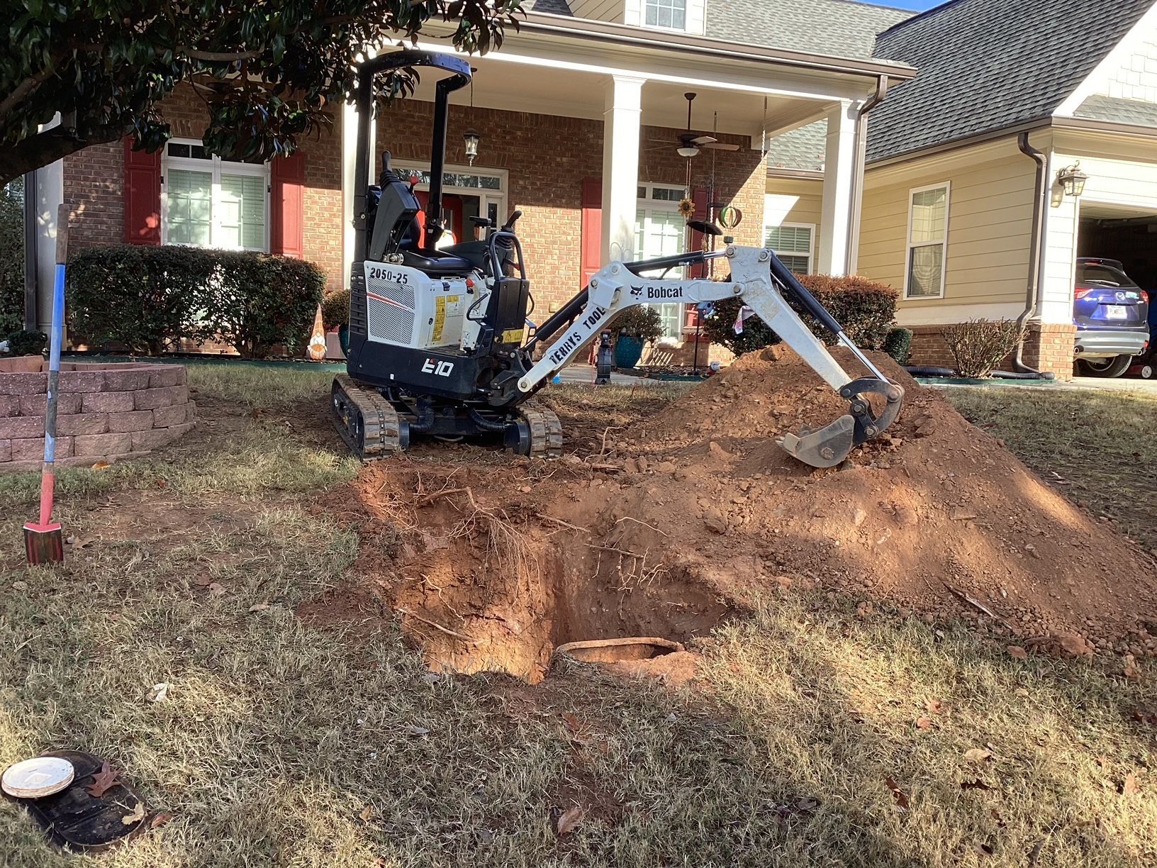 A small excavator is digging a hole in the grass in front of a house.