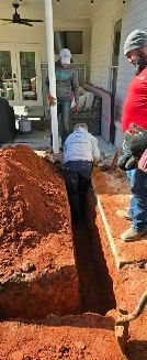 Three construction workers dig a narrow trench alongside a porch near a red dirt mound.