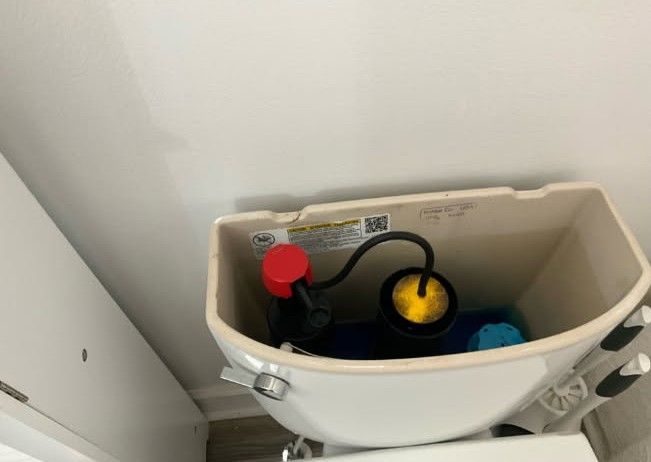 Open toilet tank with red and yellow float valves, and black tube against a white wall.