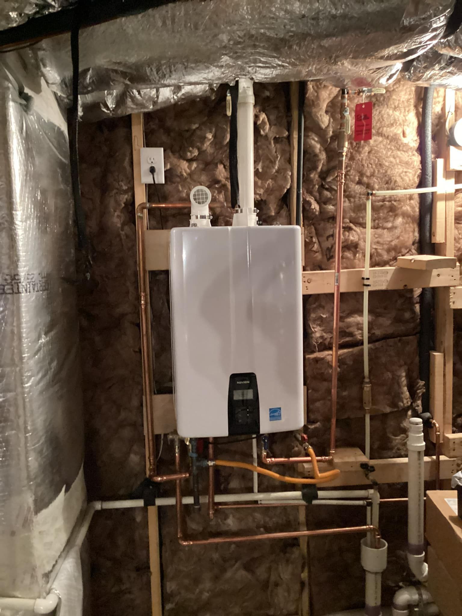 White tankless water heater mounted on a wooden frame, surrounded by copper pipes and insulation.