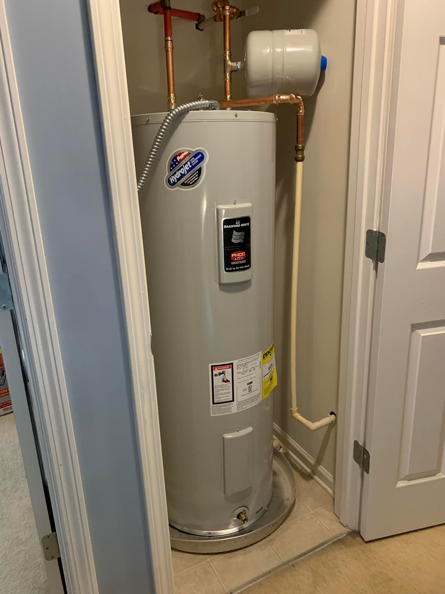 A tall, silver electric water heater in a closet with copper pipes and a white expansion tank.