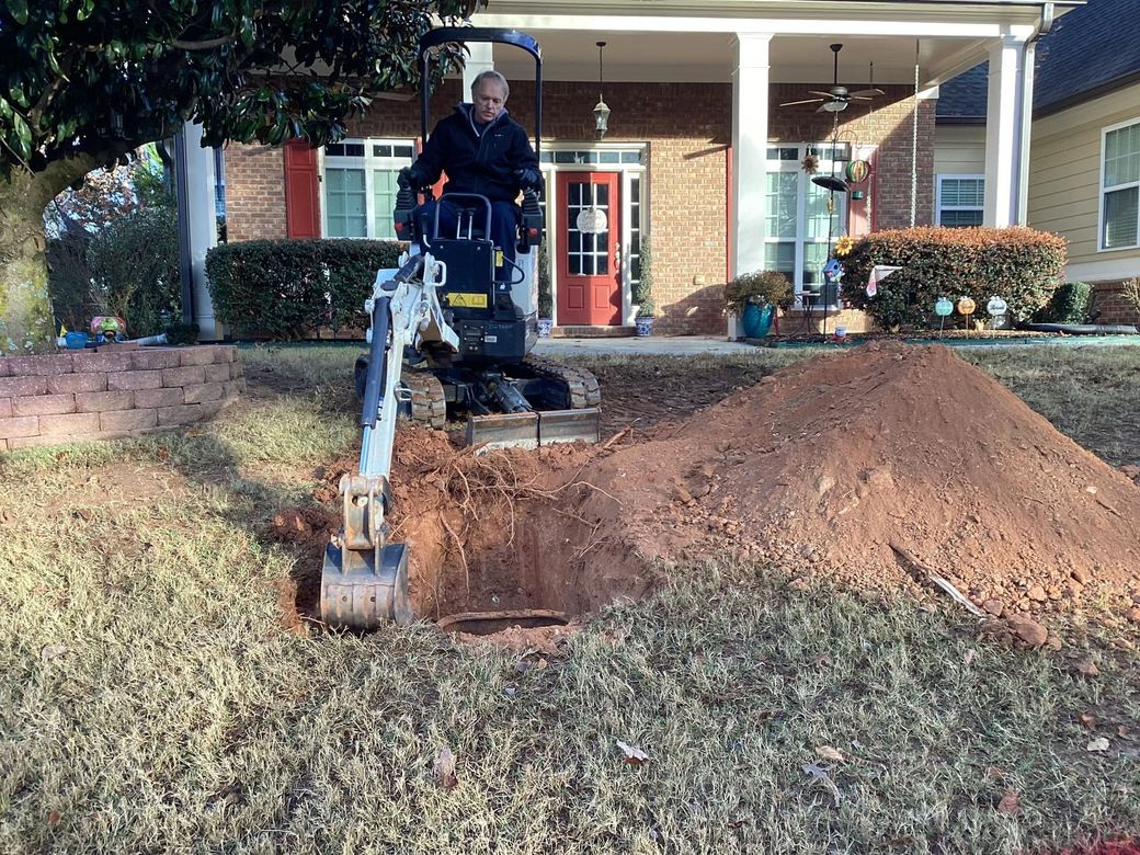 Man operating mini excavator, digging a hole in front yard of a brick house with a red door.