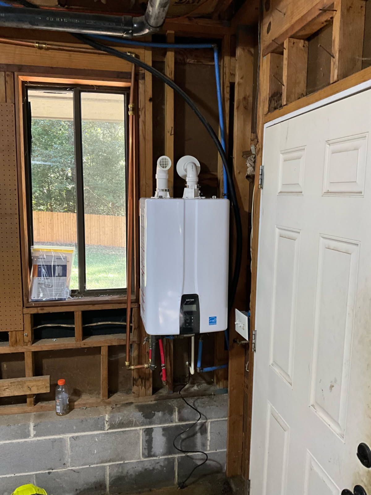 Gas water heater mounted on a wall in a room under construction, with visible pipes and unfinished walls.