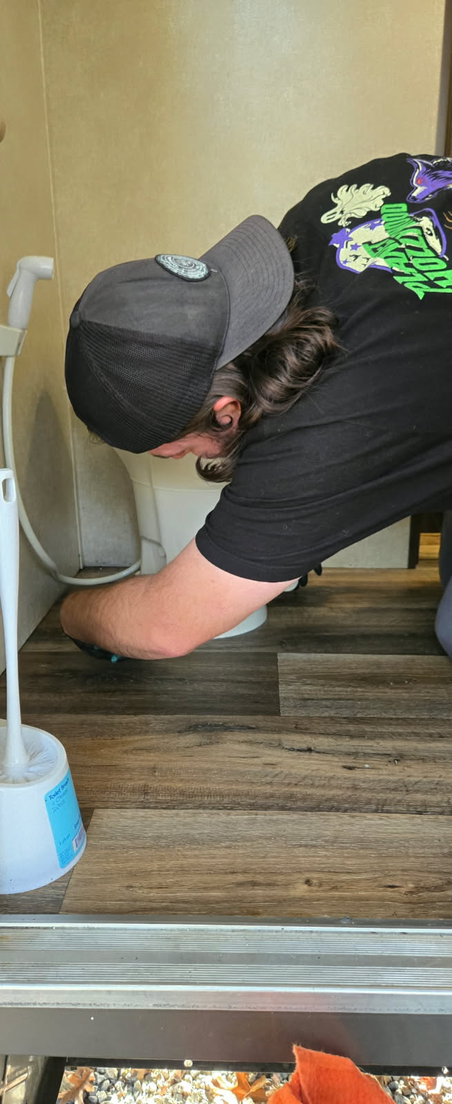Person in a cap working on a toilet, wood-like floor, white walls.