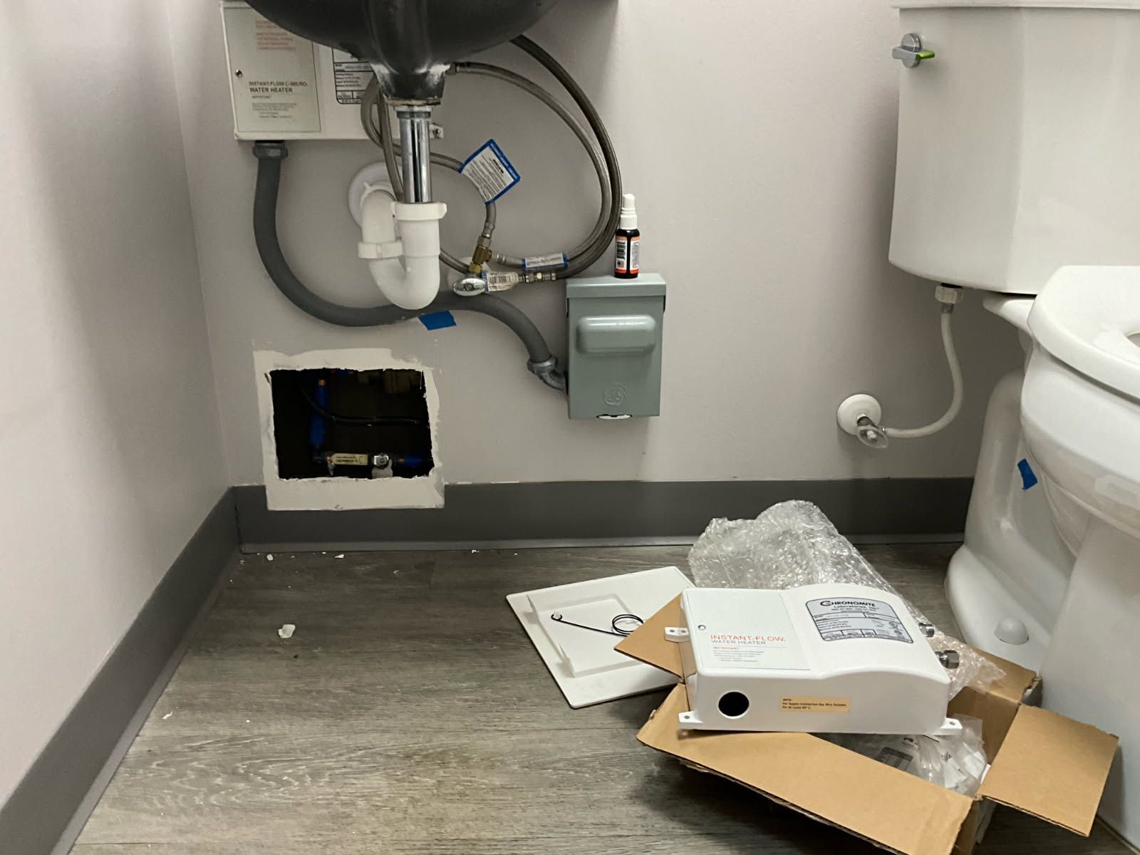 Bathroom with a sink, toilet, and exposed plumbing. A device and open box sit on the floor.