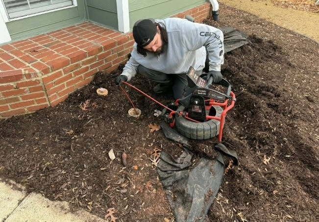 Man using a camera inspection device on a buried drain pipe in mulch near a house.