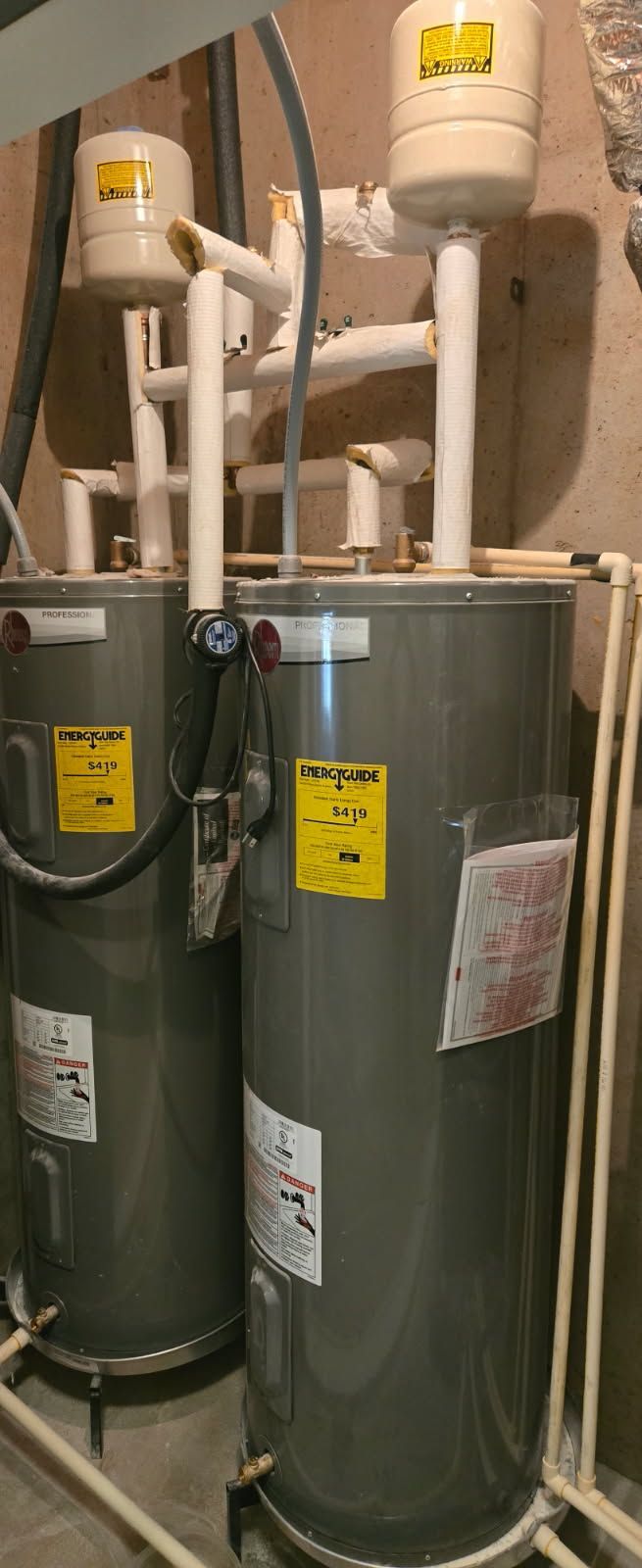 Two water heaters in a utility room. Gray tanks with white pipes and expansion tanks. Yellow energy guide stickers.