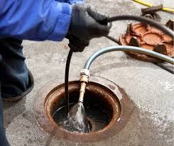 Person in blue jacket using a hose to clean a manhole; black gloves.