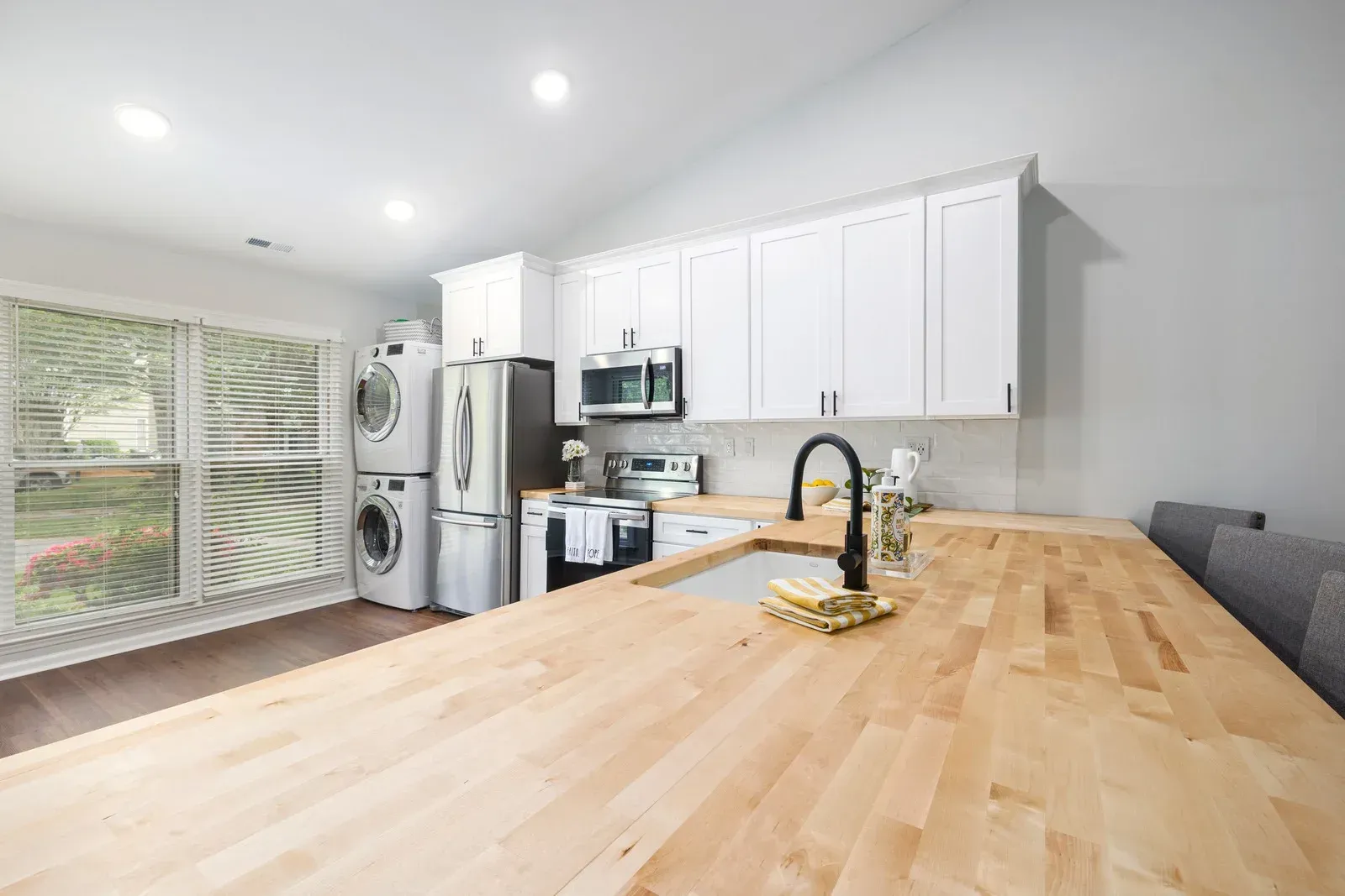 Kitchen with butcher block countertop, white cabinets, and stacked washer and dryer next to a window.