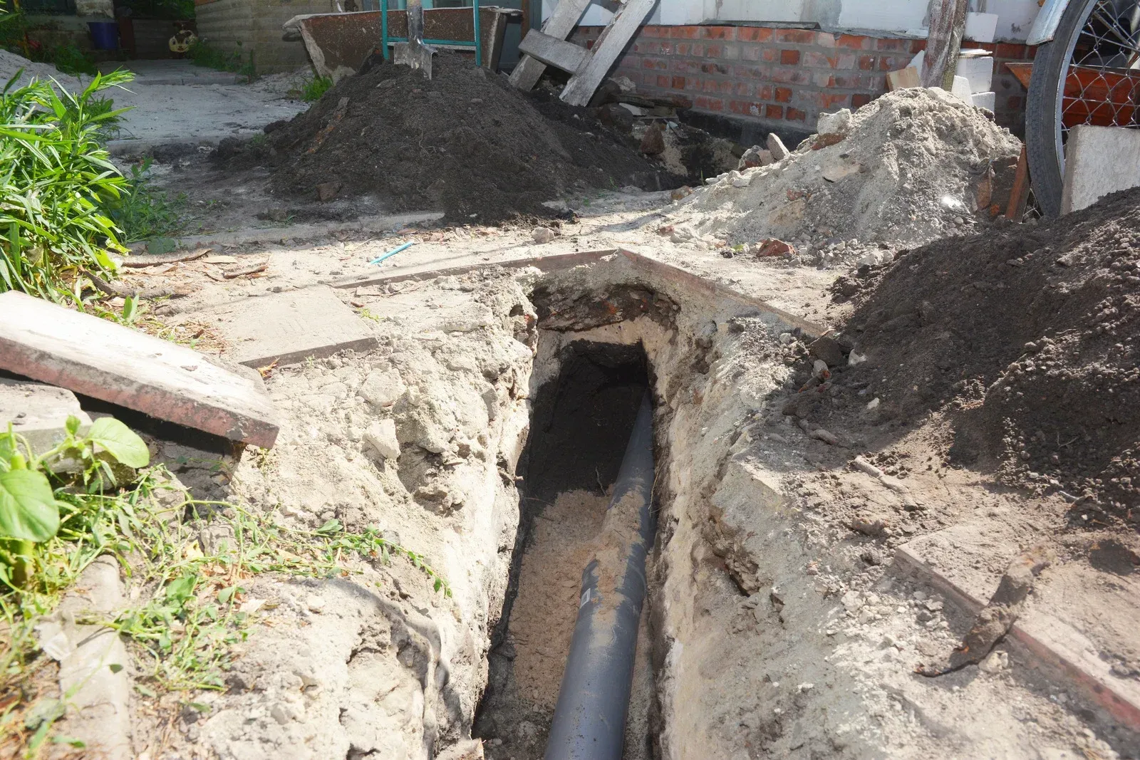 Trench dug in the ground reveals a gray pipe. Piles of dirt and debris surround the hole.