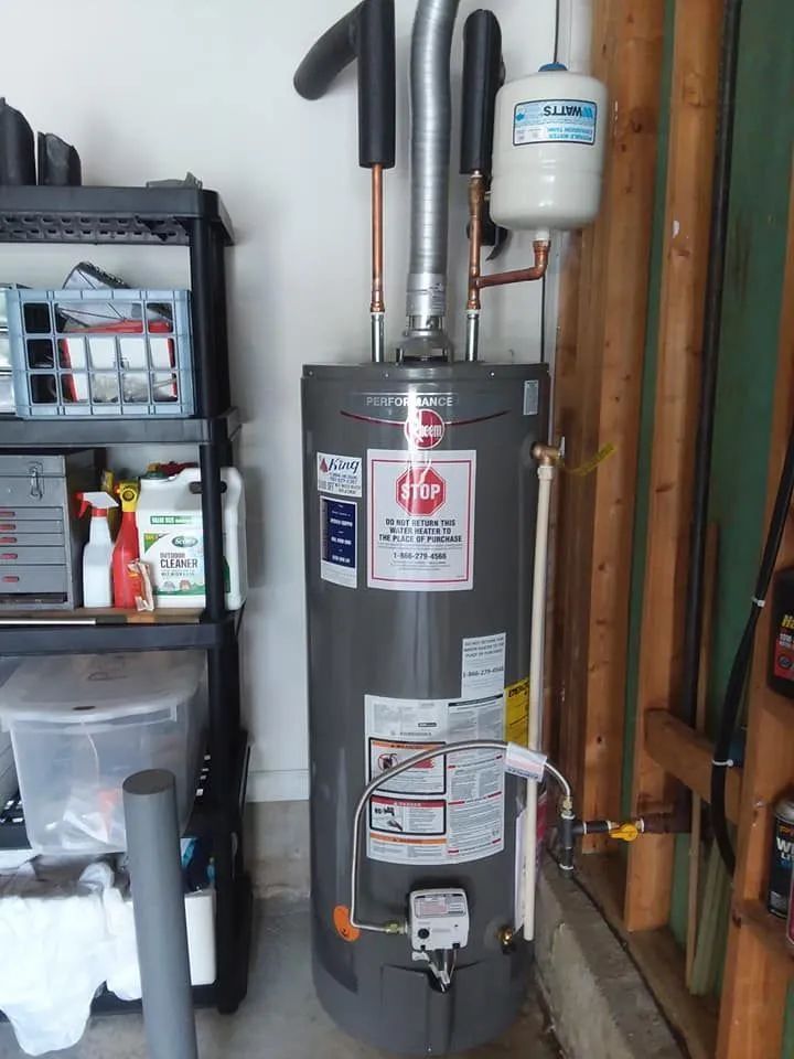 Gray water heater in a utility room with pipes, a control box, and storage shelves nearby