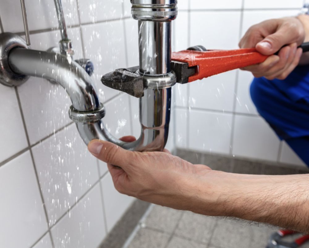Plumber using a wrench to repair a leaking sink pipe in a tiled bathroom.