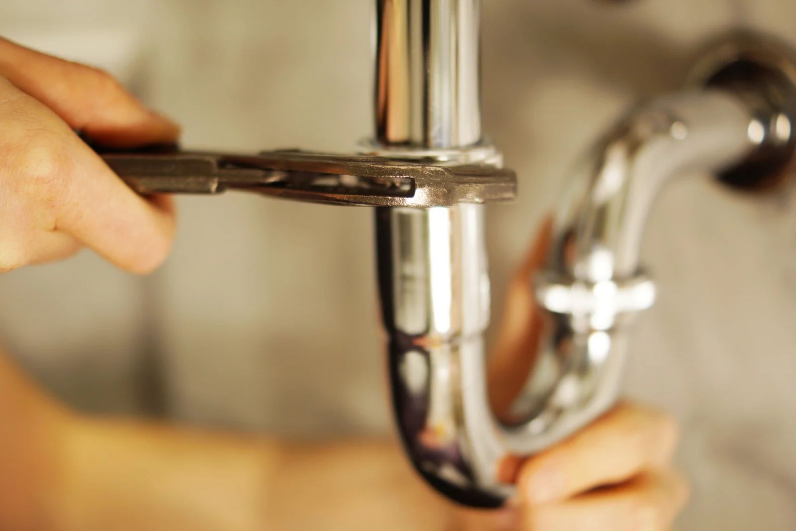 Hands using a wrench to tighten a chrome sink pipe under a faucet