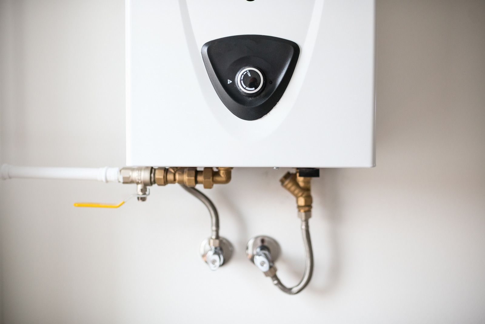 Wall-mounted white water heater with exposed brass pipes and valves against a light wall