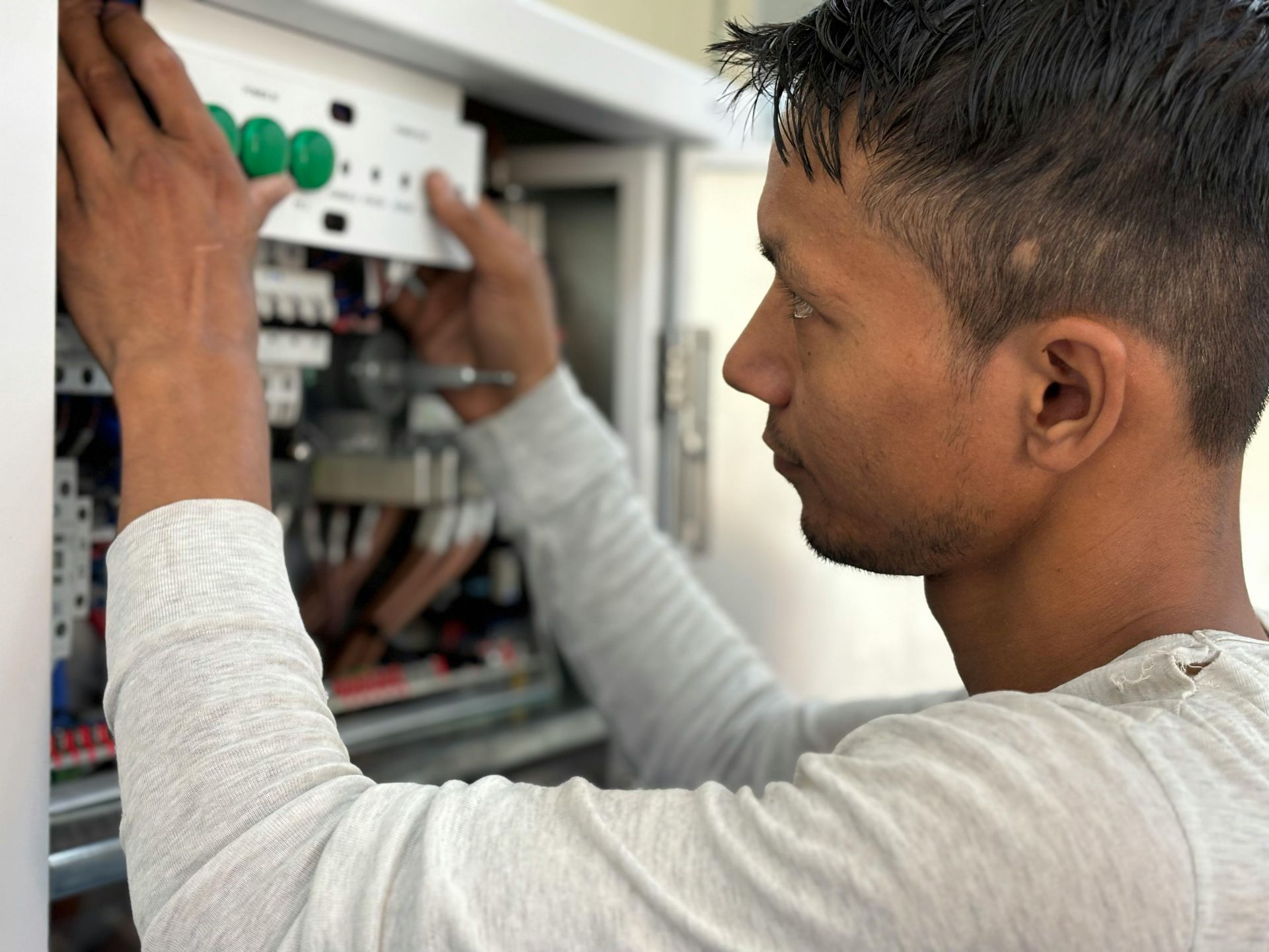 Man working on electrical panel, adjusting components inside, wearing a gray shirt, in a well-lit space.