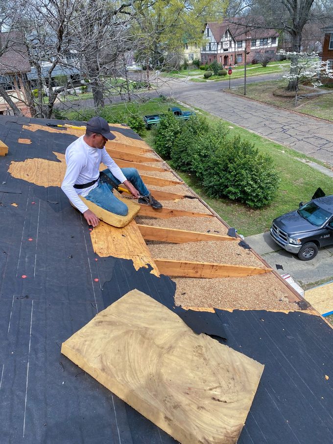 Roofer on a roof, removing shingles. He wears a hat and safety harness. Road and houses are in the background.