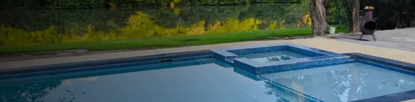Outdoor backyard swimming pool with blue water, stone coping, and trees and greenery in the background