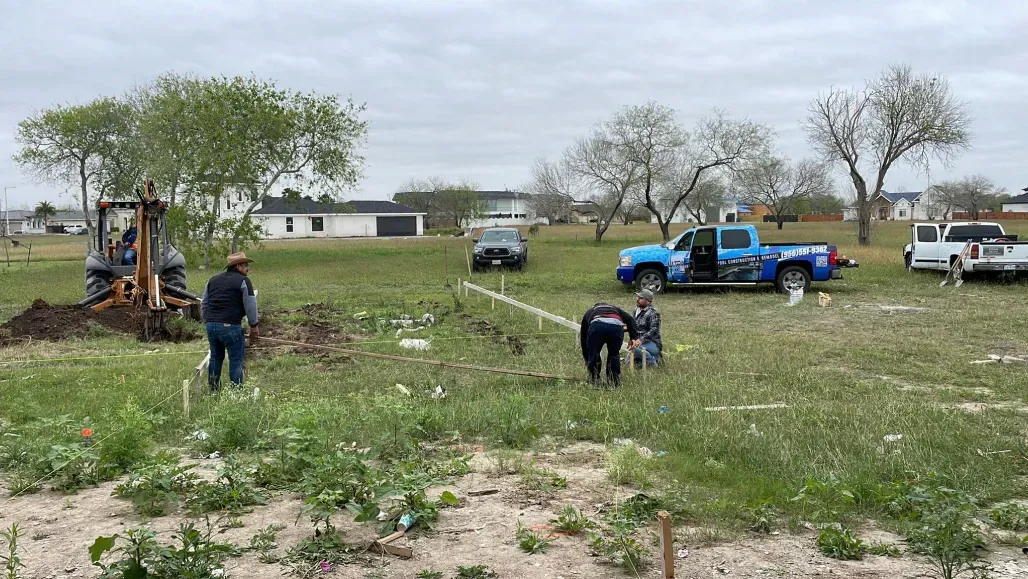 Workers clearing brush in a grassy lot with trucks and an excavator under cloudy skies